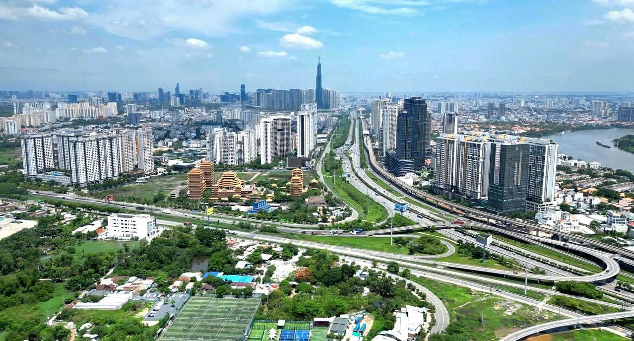 Aerial view over eastern Ho Chi Minh City: the Hanoi Highway corridor, the Metro Line 1 viaduct and the Cat Lai interchange, with the city centre skyline on the horizon