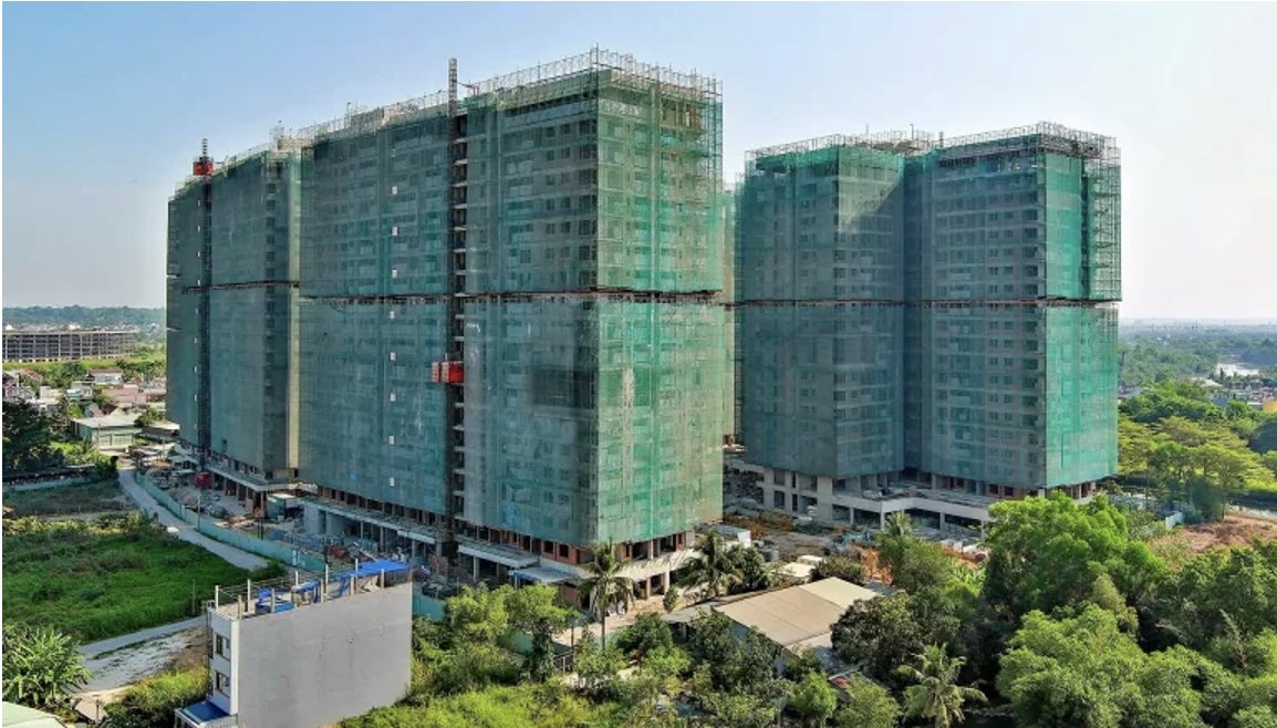 Aerial site photograph of Elysian in March 2026: the towers topped out under green safety netting with scaffolding at roof level, the podium and ground floor taking shape below, and the surrounding low-rise neighbourhood and greenery around the site