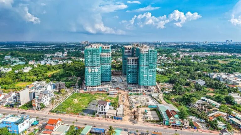 Aerial site photograph of Elysian in June 2026: the four towers topped out, safety netting coming off the upper floors and facade work underway, with Lo Lu Road in the foreground