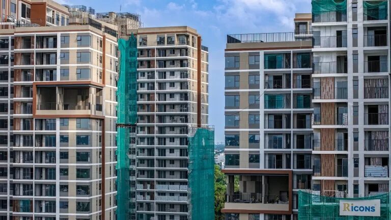 Close aerial photograph of Elysian in August 2026: facade cladding, glazing and balconies largely complete on the towers, with the last sections of green safety netting still in place